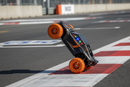 Ultra-realistic video of a black and orange toy stunt car with large textured off-road orange wheels, accurate proportions, stable geometry, performing a two-wheel tilt stunt on a professional race track. The car drives forward balanced on only two wheels, maintaining perfect shape with no distortion or warping. Inside the driver’s cabin, colorful LED lights flicker rapidly, glowing through the windshield (no lights on wheels or exterior). Dynamic camera movement: slow tracking shot from the side, then a lo